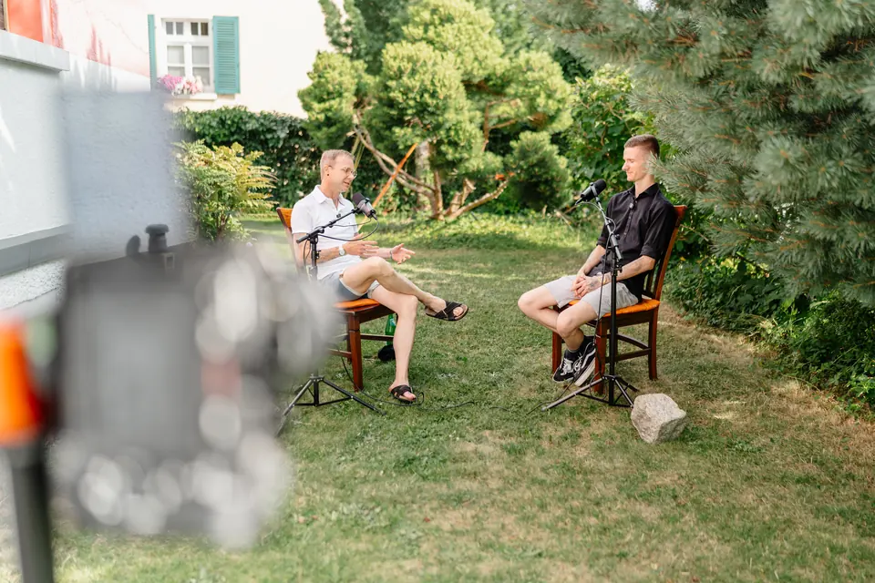 Zwei Gesprächspartner mit Mikrofonen bei einer Video-Podcast-Aufnahme im Garten, Kamera im Vordergrund