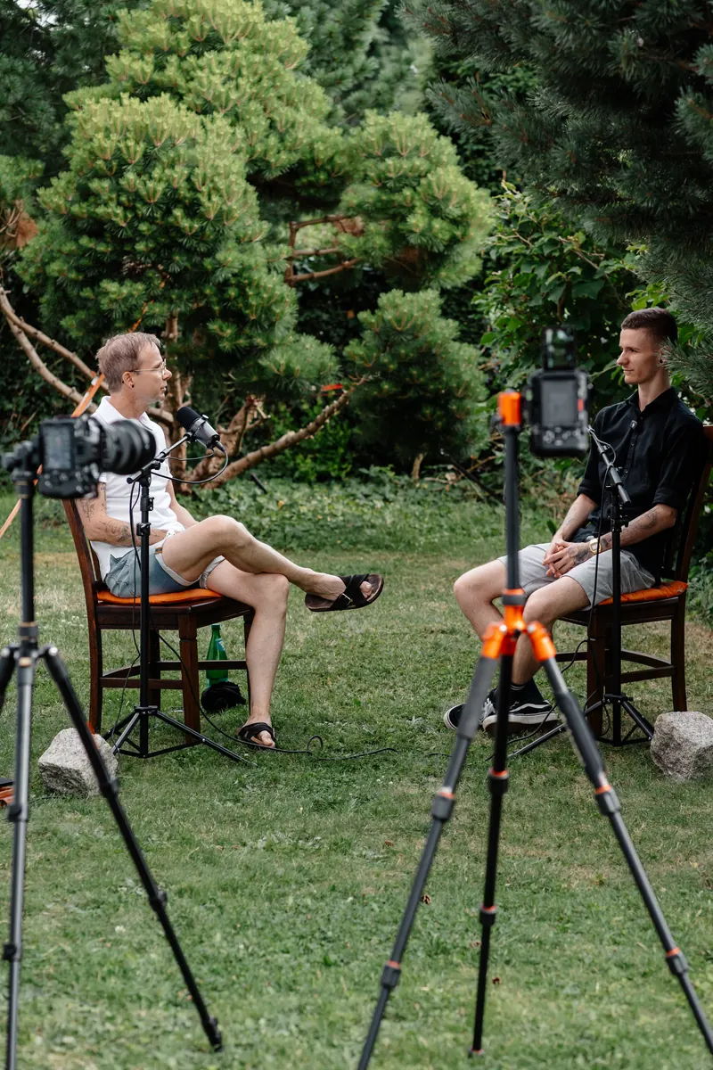 Zwei Gesprächspartner mit Mikrofonen und Kameras bei einer Podcastaufnahme im Garten